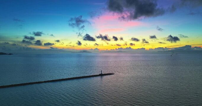 Aerial view of a long breakwater extending into calm blue ocean waters under a vibrant sunset sky filled with soft colorful clouds.