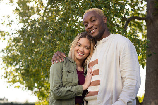 African American couple standing close together in park by tree trunk, smiling with layered jackets
