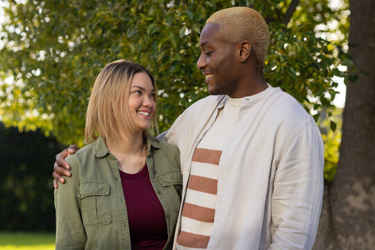 African American couple embracing beneath leafy tree in park, khaki jacket and necklace showing