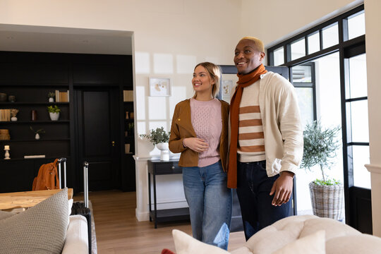 Couple standing in casual clothes near glass-paned entrance touring living room with suitcase