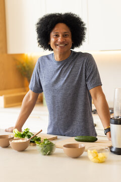 African American man in grey tee standing at kitchen counter prepping with board cucumber blender
