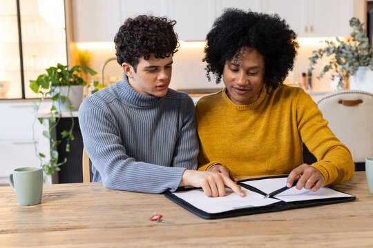 African American males in sweaters reviewing black binder with pen at kitchen table with green mugs