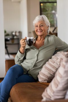 Senior woman in green blouse sitting on brown leather sofa at home holding glass mug smiling