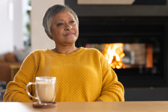 Senior African woman sitting at home in mustard-knit sweater gazing at fireplace with latte-mug