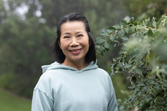 Asian senior woman standing and smiling in park wearing mint hoodie near leafy shrub buds