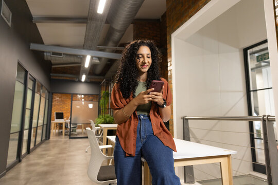 African American woman sitting on light wood table in modern co-working space holding pink phone
