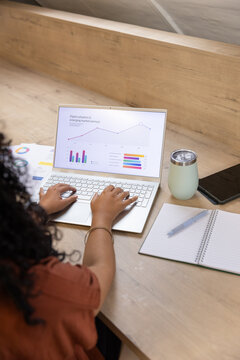 African American female working on laptop displaying charts at loft nook with notebook, tumbler