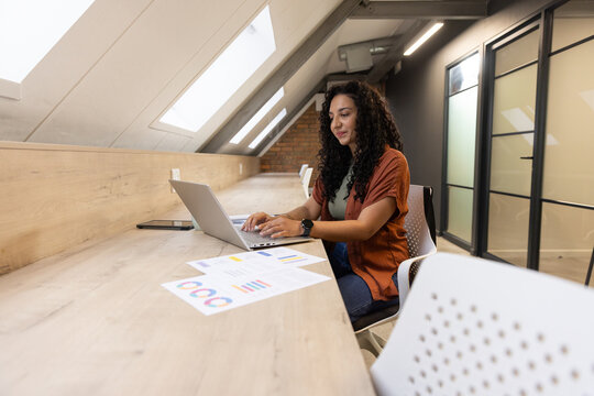 Woman working on silver laptop at loft wearing rust-orange overshirt, charts, smartphone