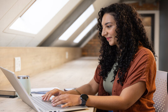 African American woman working on laptop at loft counter wearing rust shirt, smartwatch, copy space