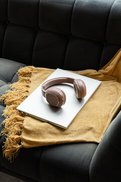 Pink-beige headphones are resting on closed silver laptop on mustard throw over dark-tufted sofa