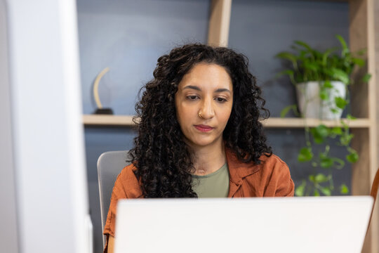 African American woman wearing rust overshirt typing on laptop at home office with shelving plant