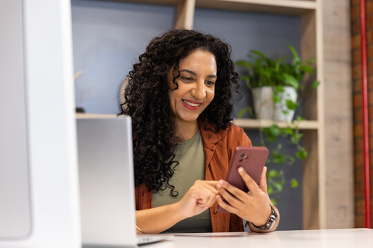 Smiling, holding maroon phone, African American woman in green top, rust jacket at desk with laptop