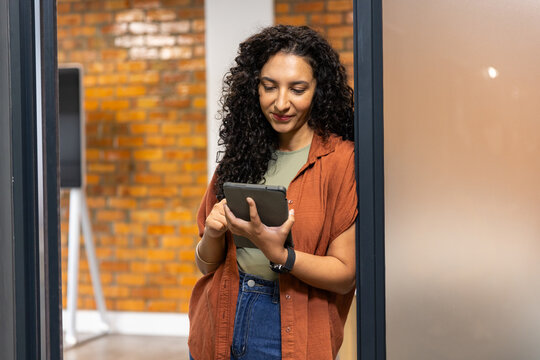 African woman standing in doorway of office wearing rust top holding tablet checking smartwatch
