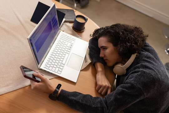 African American man resting at table at home holding phone beside laptop and headphones