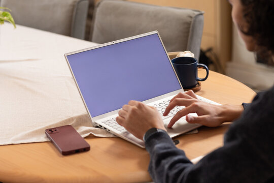 African American man typing on white laptop at home table in dark sweater, maroon-phone, copy space