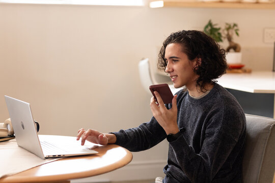 Man sitting at round table at home speaking into smartphone while using laptop with headset