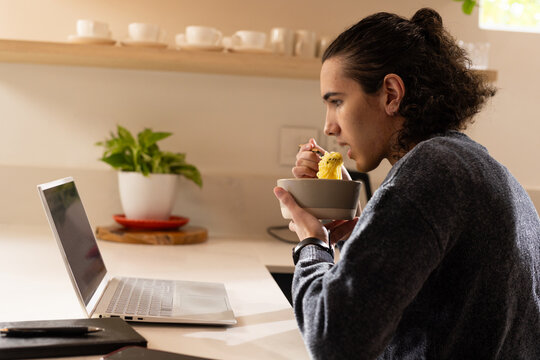 African American man sitting at kitchen counter eating from bowl with fork while viewing laptop
