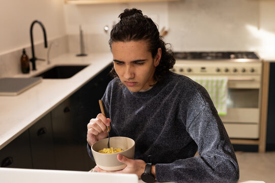 Adult male sitting at kitchen counter holding beige bowl and gold fork while viewing laptop