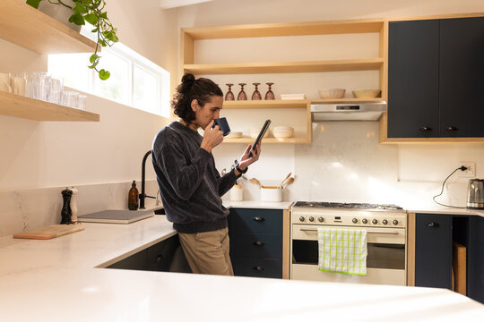 Adult African American man leaning on quartz countertop in modern kitchen, holding tablet and mug