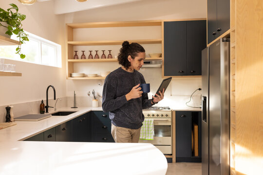 Adult male standing in home kitchen wearing charcoal sweater holding tablet and navy mug by oven