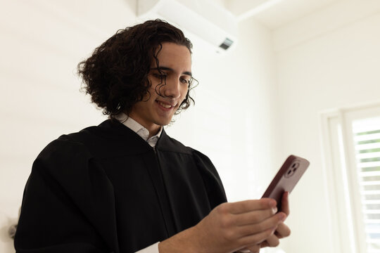 African American man in black gown white collar holding red phone looking down smiling at home