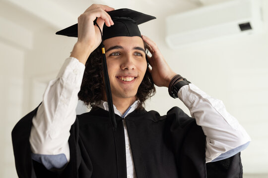 Man in black graduation gown adjusting mortarboard cap and smiling, showing bracelets in pale room