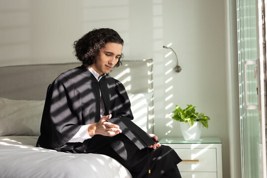 African American man wearing graduation gown sitting on bed in bright bedroom holding mortarboard