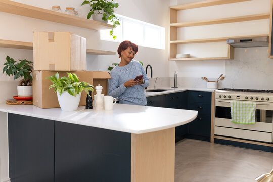African American woman wearing blue sweater, holding smartphone, unpacking moving boxes in kitchen