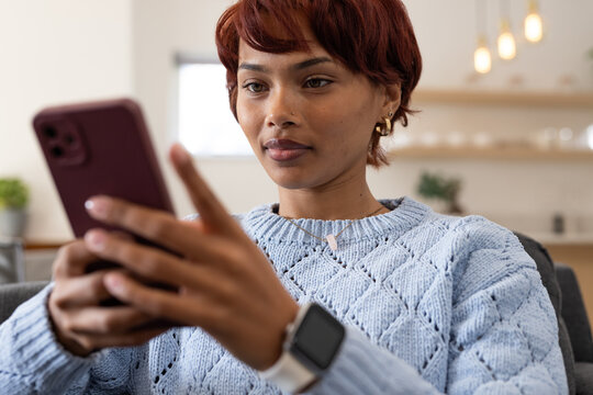 Adult African American woman wearing blue sweater and smartwatch holding maroon phone on gray couch