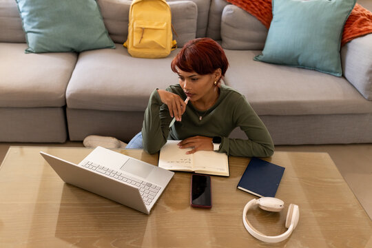 African American woman leaning on low coffee table at home, holding pen over notebook, using laptop