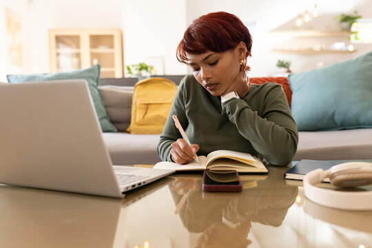 African American woman wearing green sweater sitting at home writing in notebook near laptop