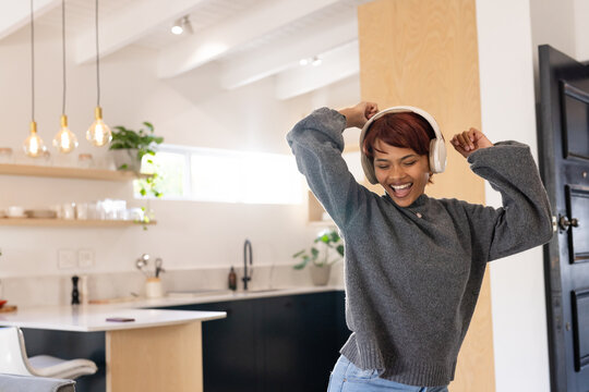 African American woman dancing in kitchen wearing white headphones with phone on counter