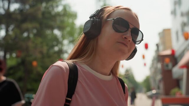 Young woman wearing sunglasses and headphones walks outdoors in public space with trees and distant people visible. Woman carries backpack while background shows urban elements