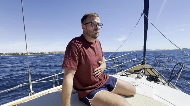 Man feeling seasick and gagging while sitting on a sailboat at sea, showing nausea, motion sickness, discomfort and travel sickness during a sailing trip.