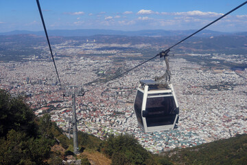 Aerial view of a white cable car cabin gliding over a vast cityscape with endless rooftops and distant mountains on a sunny day © Альфира Зиганшина
