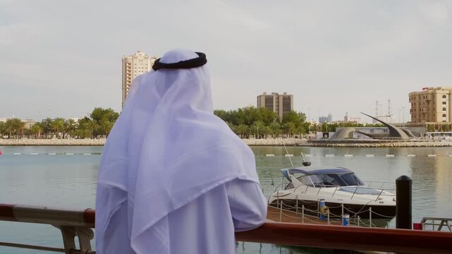 Rear view of man wearing traditional white kandura, ghutra, and agal observing boats moored in calm harbor waters with modern cityscape beyond.