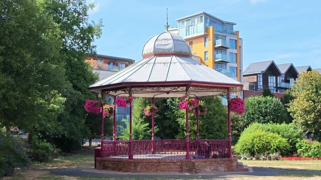 Newbury Berkshire England  UK. 04.09.2026. Video. A picturesque park scene featuring a classic bandstand adorned with hanging flower baskets, set against a backdrop of contemporary urban architecture