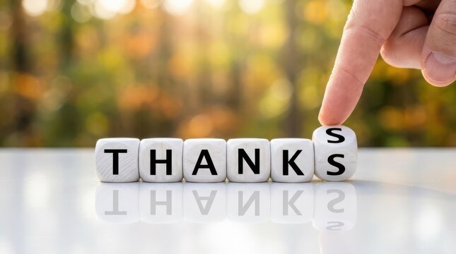 A hand placing a wooden cube with the letter s to complete the word thanks on a reflective surface with a blurred nature background