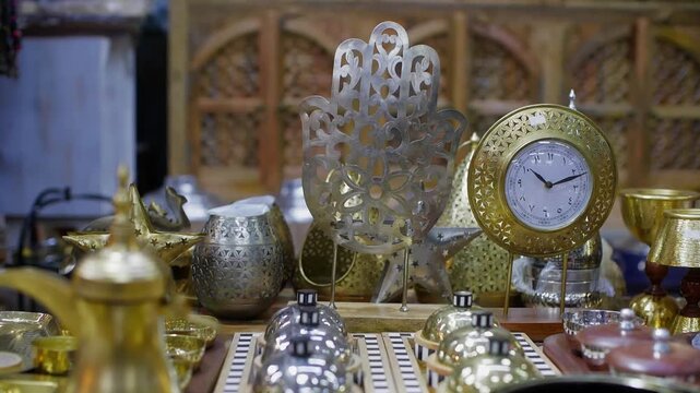 Intricate brass Islamic artifacts arranged on table, featuring decorative clock with ornamental filigree patterns, metalwork vessels, glasses, symbolic home decor items.