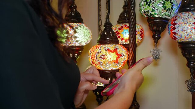 Shopper inspects illuminated Turkish style lantern with intricate colored glass mosaic pattern, brass metalwork, hanging in retail display with similar decorative lamps.