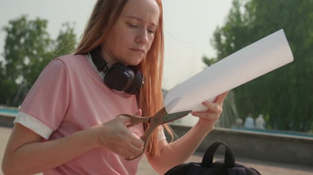 Young woman with long hair cuts sheet of paper outdoors wearing pink shirt and headphones around her neck. Woman holds cut paper in one hand and scissors in other against urban park backdrop