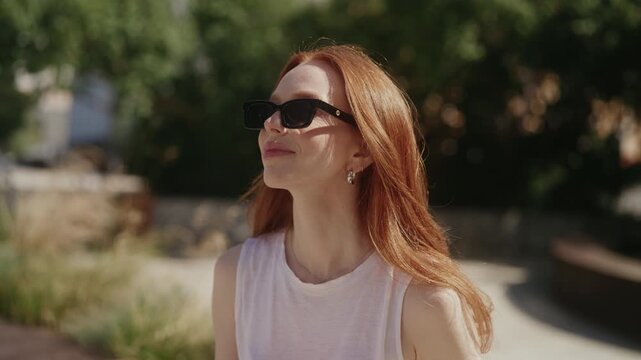 Portrait of young gorgeous woman with ginger hair wearing sunglasses enjoying summer weather and drinking fresh mojito cocktail outdoors in park on beautiful sunny day. Shooting in slow motion.