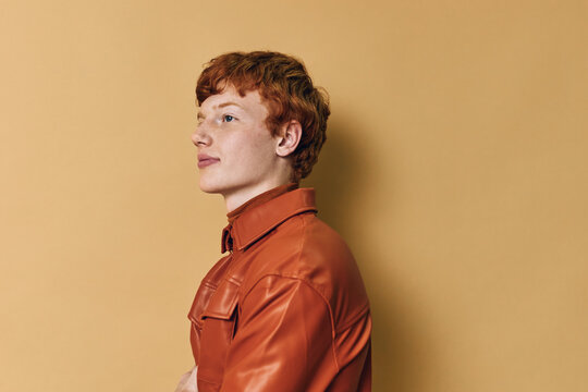 Portrait of young redhead man wearing orange leather jacket standing sideways on beige background with confident expression and relaxed posture in studio light.