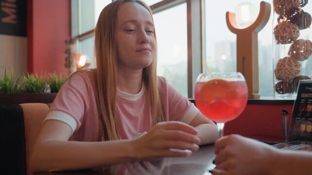 Young woman sits at table in indoor setting engaging with another woman across table. Woman receives and drinks from large red cocktail glass garnished with orange slice during their