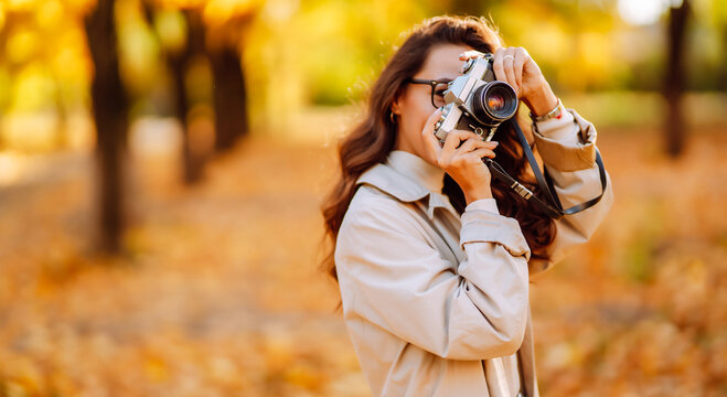 Cheerful woman sits in park surrounded by colorful autumn leaves, holding camera. Woman in coat relaxes in autumn park and takes photos at sunset. Concept of leisure, beauty, and enjoyment.
