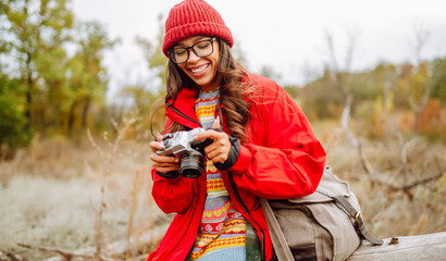 Young woman in bright jacket stands with camera in an autumn forest. The traveler in the red jacket enjoys the vibrant colors of autumn outdoors. A concept of leisure and walking. © maxbelchenko