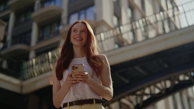 Low-angle view of stunning young woman with long red hair smiling, looking happy using mobile phone for texting, browsing, scrolling on sunlit city street with buildings in background, slow motion.