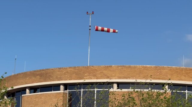 A red and white striped windsock flutters horizontally on a tall metal pole, fully extended by strong wind against a clear blue sky.
