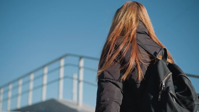 Woman with long hair walks away from camera wearing black jacket and carrying backpack. scene is set outdoors under clear blue sky with metal railings in background showing consistent