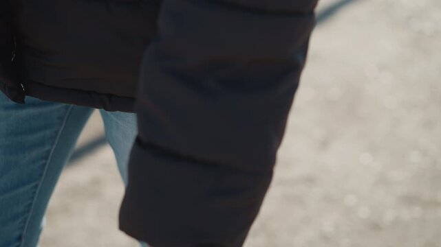 Woman walks outdoors wearing black jacket and blue jeans. focus remains on her lower body as woman moves along paved path with natural light casting shadows from nearby objects